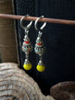 A pair of handmade dangle earrings hanging from a wooden rod. The earrings feature silver-toned metal hoops and beaded details with turquoise, red, and yellow beads, set against a dark textured background with rustic wood props.